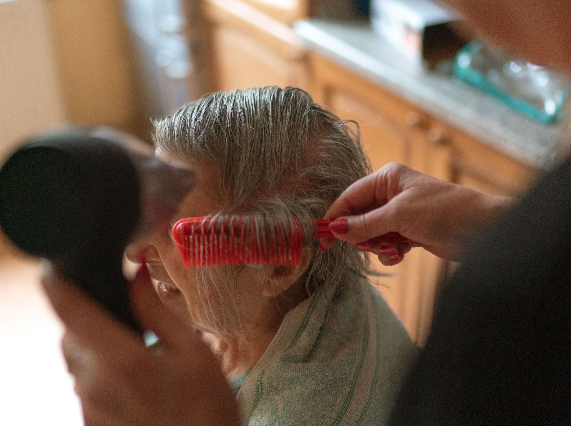 A caregiver assists a senior adult with hair drying and combing at home.